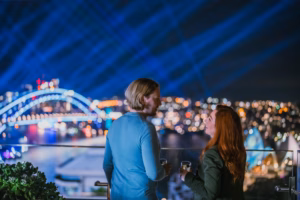 Two people standing on a balcony overlooking the sydney harbour bridge.