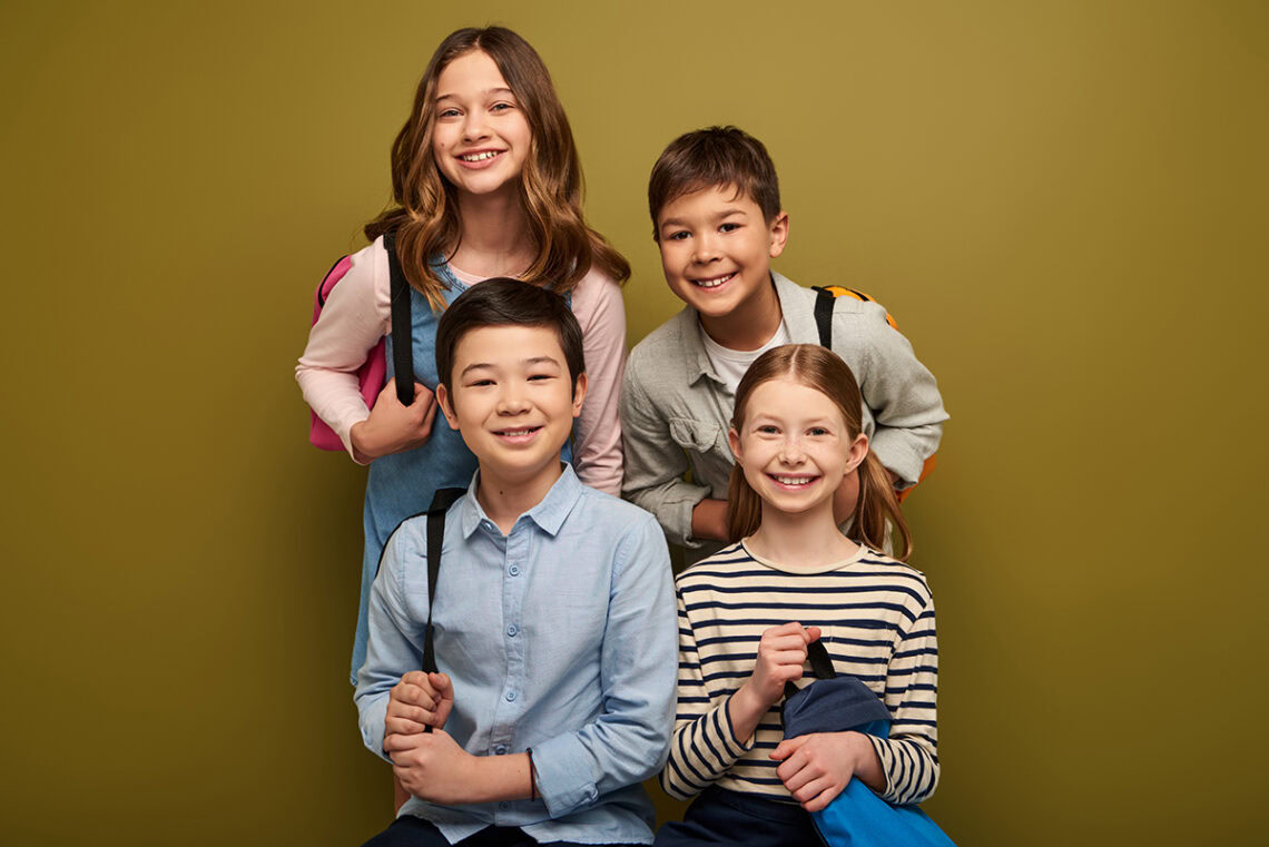 Smiling multiethnic schoolkids with backpacks looking at camera near friends on khaki background