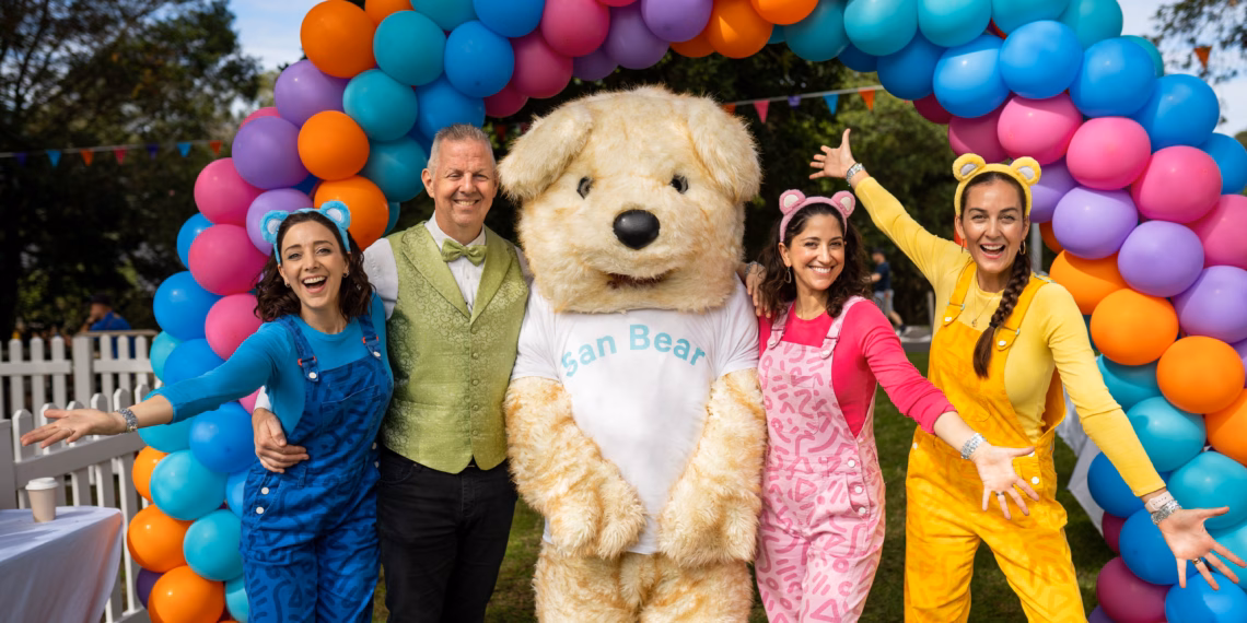 Three people in colorful outfits pose with a large teddy bear.
