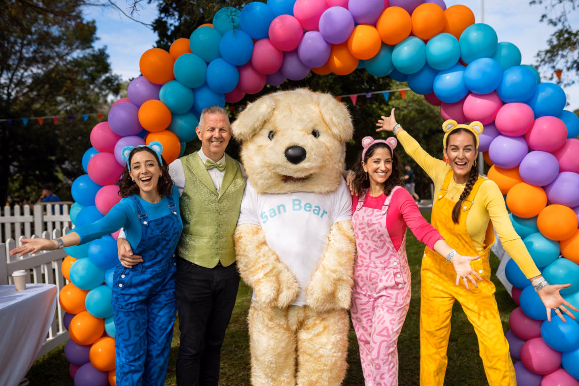 Three people in colorful outfits pose with a large teddy bear.