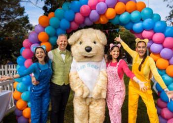 Three people in colorful outfits pose with a large teddy bear.