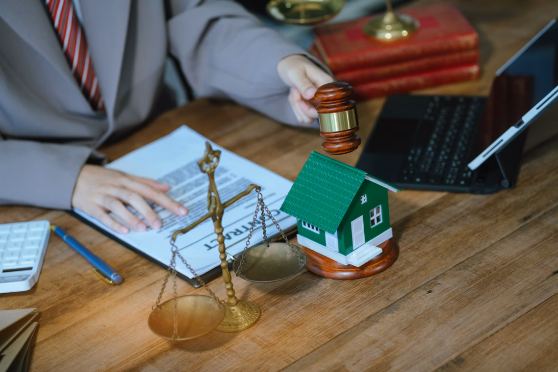 A judge's gavel is holding a house model and scales of justice on a wooden table.