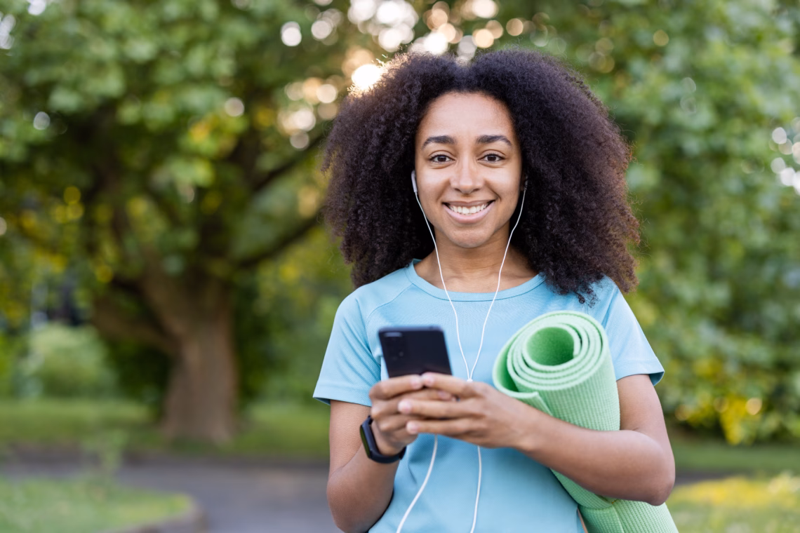 Woman smiling with a yoga mat and smartphone, ready for an outdoor exercise session in a green park setting.