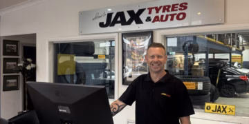 A man wearing a black shirt with a "JAX" logo on it stands behind a counter at a JAX Tyres & Auto shop. The counter is glass, and on it are a computer monitor, keyboard, and various promotional materials. To the right of the man, a sign reads "Put a Cap on Breast Cancer!" with a pink ribbon logo. Behind the man, a large vertical sign with red lettering says "JAX TYRES & AUTO." In the background, tires and other auto parts are visible.