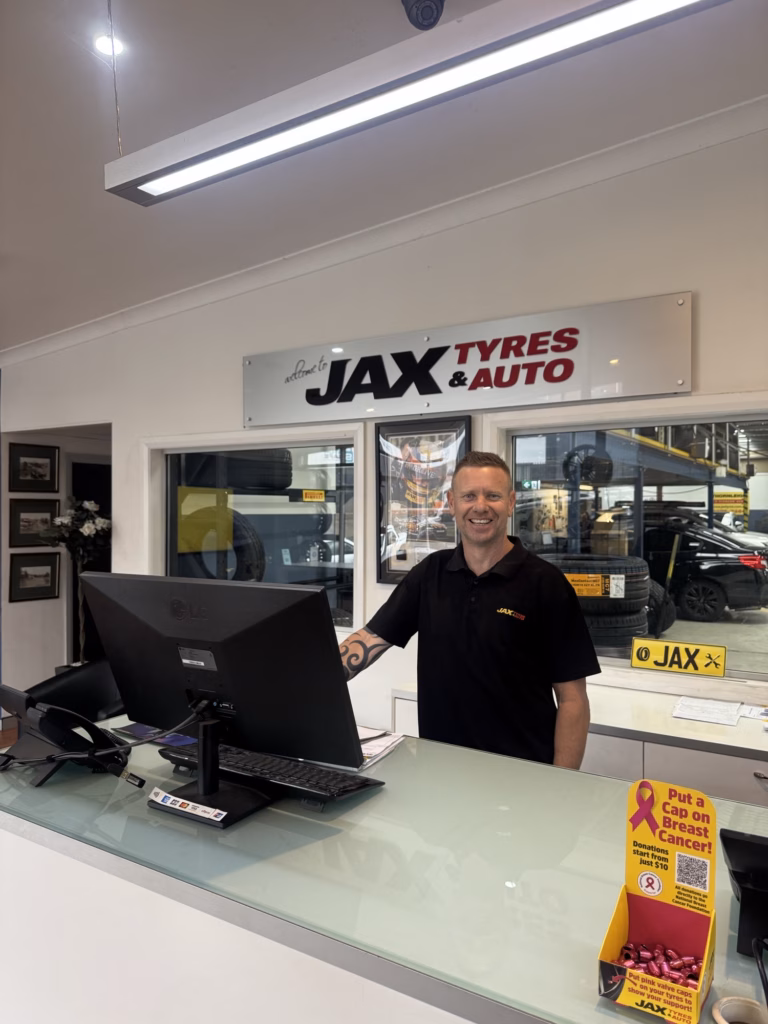 A man wearing a black shirt with a "JAX" logo on it stands behind a counter at a JAX Tyres & Auto shop. The counter is glass, and on it are a computer monitor, keyboard, and various promotional materials. To the right of the man, a sign reads "Put a Cap on Breast Cancer!" with a pink ribbon logo. Behind the man, a large vertical sign with red lettering says "JAX TYRES & AUTO." In the background, tires and other auto parts are visible.