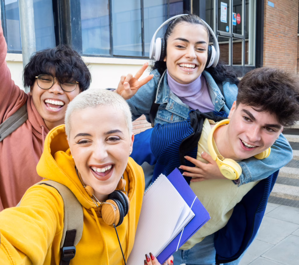 A diverse group of four happy students taking a selfie and holding notebooks, celebrating their academic success and school results.