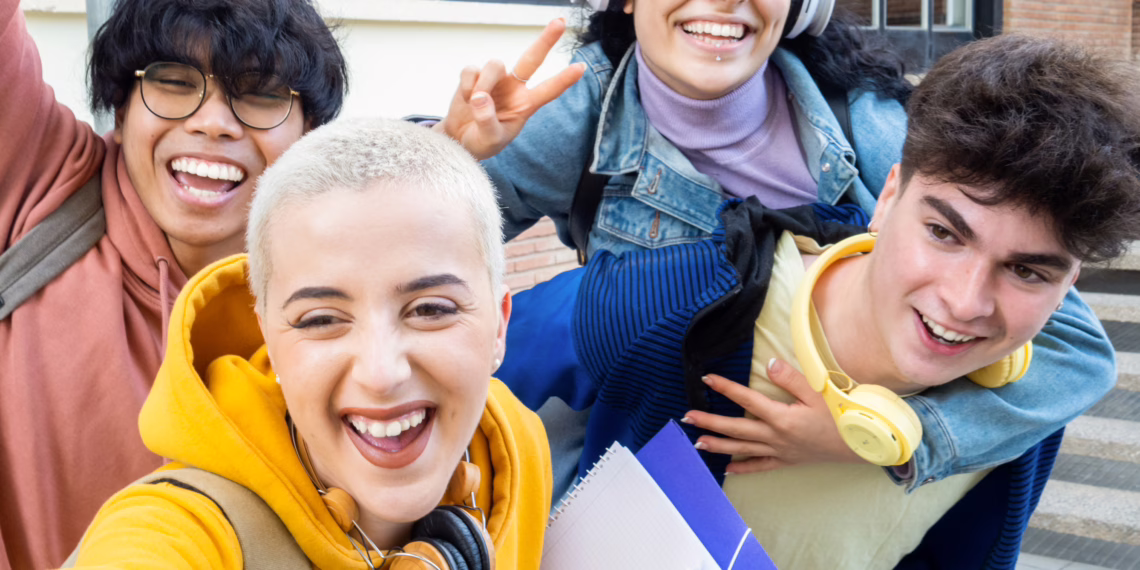 A diverse group of four happy students taking a selfie and holding notebooks, celebrating their academic success and school results.