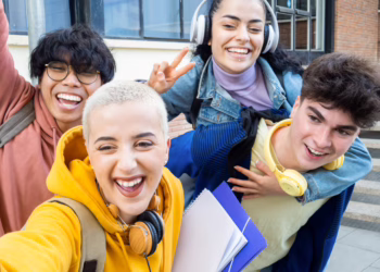 A diverse group of four happy students taking a selfie and holding notebooks, celebrating their academic success and school results.