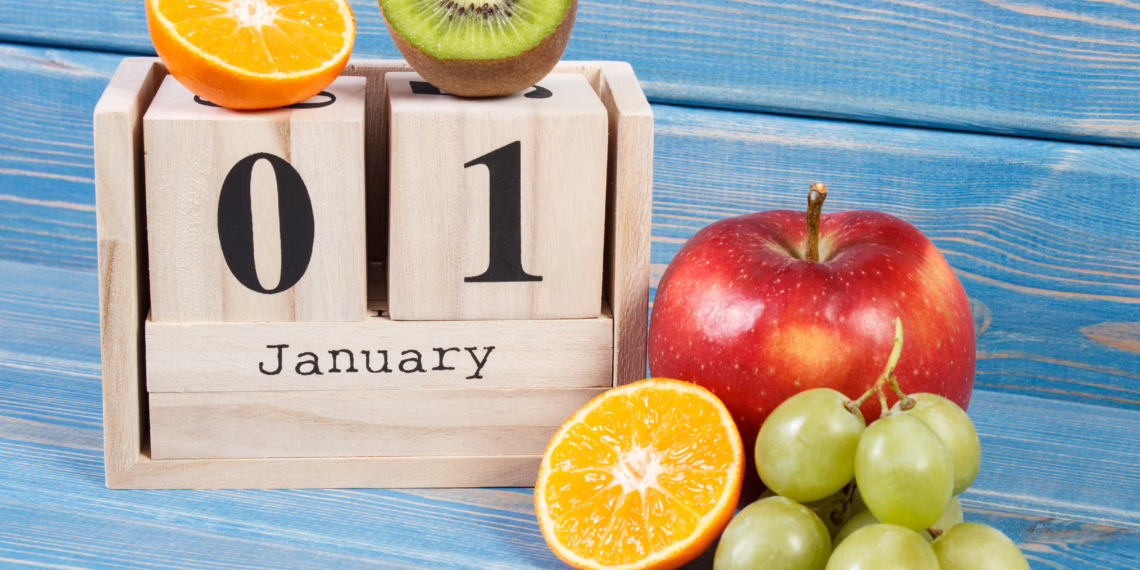A wooden block calendar displaying "01 January" sitting on a blue wooden table, surrounded by fresh fruit including a red apple, green grapes, and sliced kiwi and orange, symbolizing a fresh start to the new year.