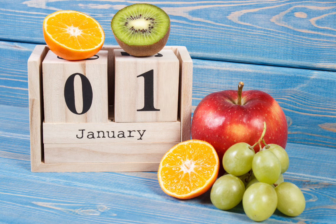 A wooden block calendar displaying "01 January" sitting on a blue wooden table, surrounded by fresh fruit including a red apple, green grapes, and sliced kiwi and orange, symbolizing a fresh start to the new year.