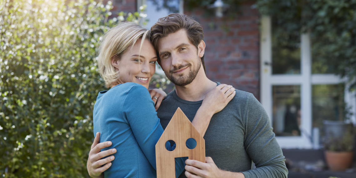 A smiling couple standing in a garden in front of a brick home, holding up a small wooden house cutout to symbolize the goal of buying a property.