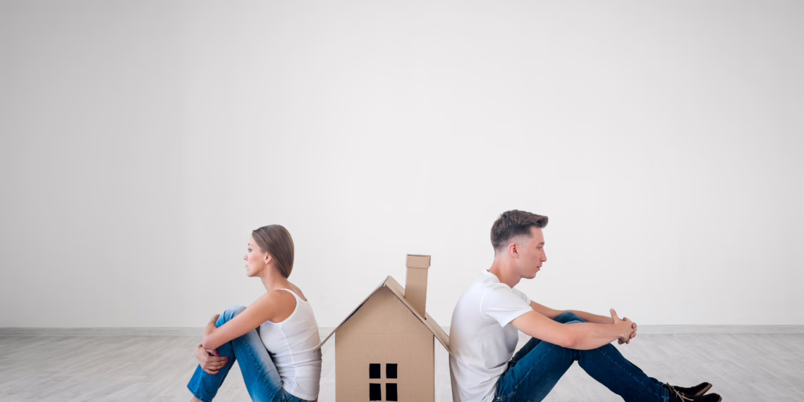 A young couple sitting back-to-back on the floor with a small cardboard house between them, looking unhappy, symbolising the challenges of dividing assets and property settlement after separation.