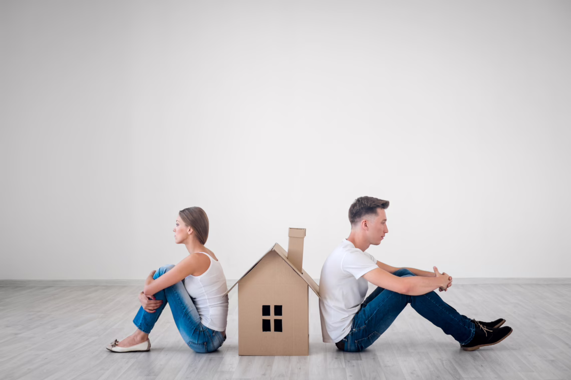 A young couple sitting back-to-back on the floor with a small cardboard house between them, looking unhappy, symbolising the challenges of dividing assets and property settlement after separation.