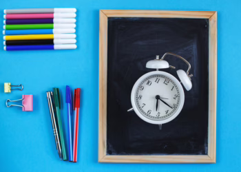 A top-down, flat lay photograph of school supplies arranged on a bright blue background. On the left side, there is a horizontal stack of colorful felt-tip markers, followed by two binder clips (yellow and pink) and four pens aligned vertically. On the right side, a white, twin-bell analog alarm clock sits in the center of a small, wooden-framed chalkboard.