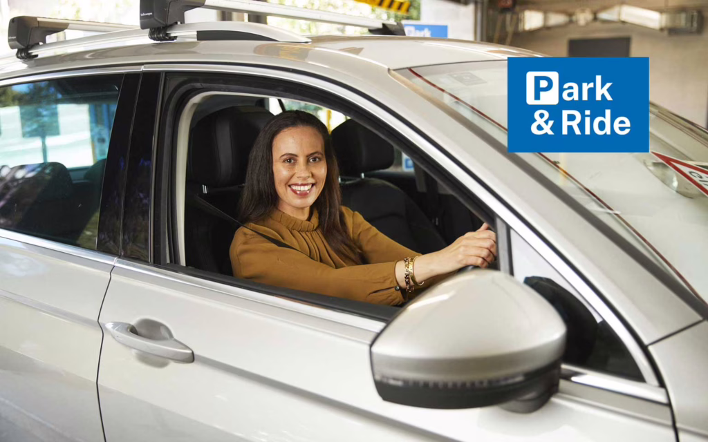 Smiling Woman entering a carpark in a silver car. The words Park & Ride in white text on a blue background. The "P" in Park is stylised like the international carpark signage.