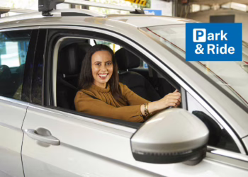 Smiling Woman entering a carpark in a silver car. The words Park & Ride in white text on a blue background. The "P" in Park is stylised like the international carpark signage.