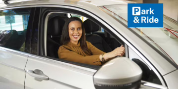 Smiling Woman entering a carpark in a silver car. The words Park & Ride in white text on a blue background. The "P" in Park is stylised like the international carpark signage.